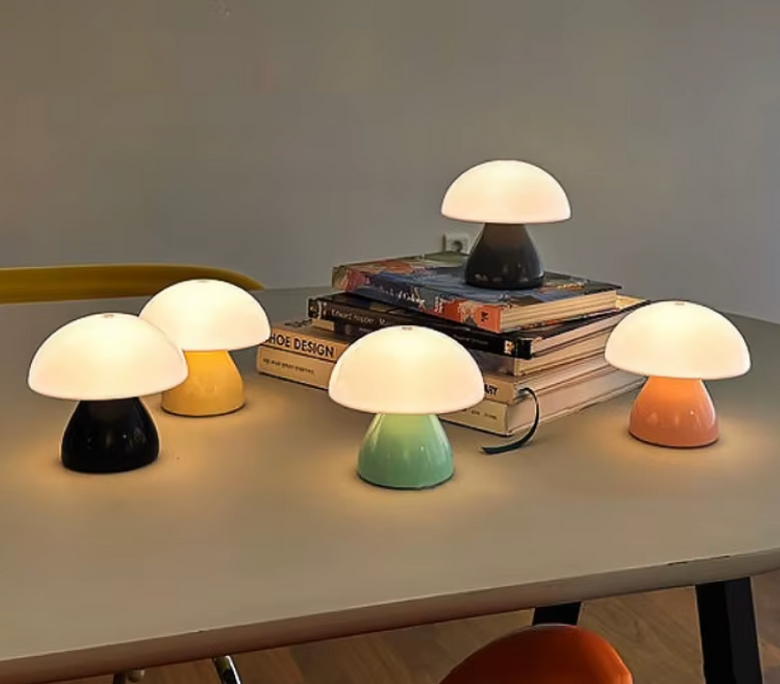 Five mushroom-shaped table lamps in different colors (black, yellow, dark gray, mint green, and peach) arranged on a tabletop with books in the background.