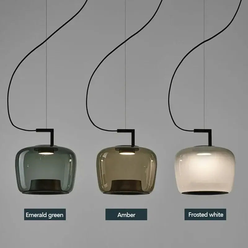 Three pendant light shades shown side by side in different color options: Emerald green, Amber, and Frosted white. Each lamp has a curved black cord and a rectangular black fixture holding a rounded glass shade.