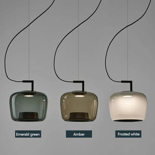 Three pendant light shades shown side by side in different color options: Emerald green, Amber, and Frosted white. Each lamp has a curved black cord and a rectangular black fixture holding a rounded glass shade.