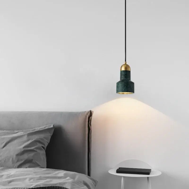 A modern pendant light with a textured dark green shade and a gold-finished brass cap, suspended over a bed in a minimalist bedroom setting.