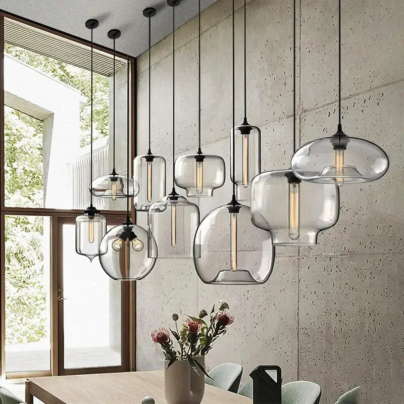 Hero A set of modern pendant lights with clear glass bell-shaped shades and black metal hardware, hanging over a dining table in a contemporary room with a concrete wall and large window.