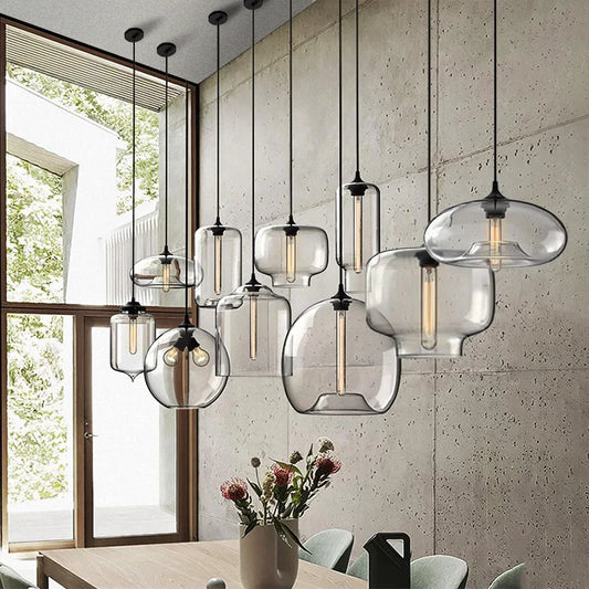 Hero A set of modern pendant lights with clear glass bell-shaped shades and black metal hardware, hanging over a dining table in a contemporary room with a concrete wall and large window.