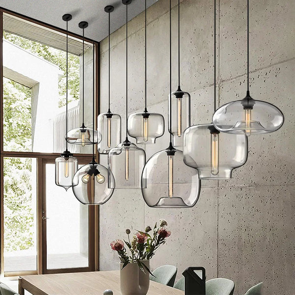 Hero A set of modern pendant lights with clear glass bell-shaped shades and black metal hardware, hanging over a dining table in a contemporary room with a concrete wall and large window.